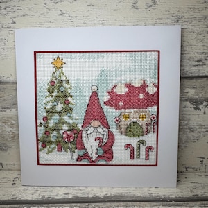 May include: A cross-stitched Christmas card featuring a gnome, Christmas tree, and mushroom house. The gnome wears a red hat and holds a candy cane. The tree is decorated with ornaments, and the mushroom house has a red and white spotted cap.