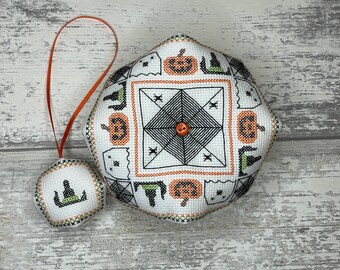 May include: Two white fabric Halloween ornaments with black and green embroidery. The larger ornament features a spiderweb design with pumpkins and ghosts. The smaller ornament features a witch's hat and a pumpkin. Both ornaments have a loop for hanging.