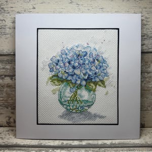 May include: A cross-stitch artwork featuring a bouquet of blue hydrangeas in a clear glass vase. The artwork is framed in black and set against a white background, mounted on a white card. The overall design is a floral theme.