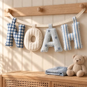 May include: A fabric banner spelling the name NOAH, featuring individual stuffed letters hanging from a beige ribbon. The N is blue gingham, the O is cream quilted fabric, the A is light blue with grey stars, and the H is blue striped.