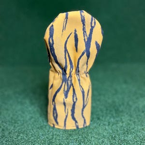Puede incluir: Una funda para la cabeza del palo de golf con un fondo amarillo y un patr&oacute;n de pinceladas azul marino. La funda es cil&iacute;ndrica con una parte superior fruncida, dise&ntilde;ada para proteger los palos de golf. La funda est&aacute; sobre una superficie verde.