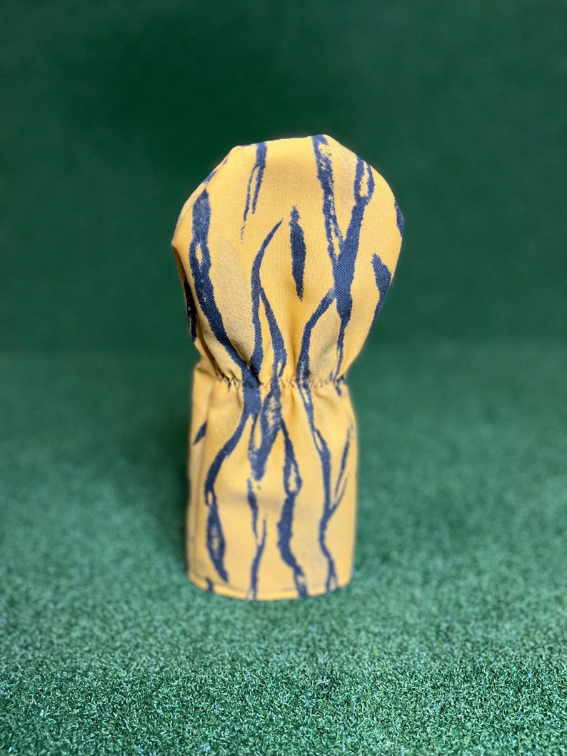 Puede incluir: Una funda para la cabeza del palo de golf con un fondo amarillo y un patr&oacute;n de pinceladas azul marino. La funda es cil&iacute;ndrica con una parte superior fruncida, dise&ntilde;ada para proteger los palos de golf. La funda est&aacute; sobre una superficie verde.