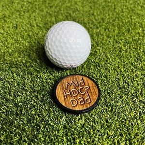 May include: A white golf ball and a wooden golf ball marker with the text "Mid HDCP Dad" are on a green surface. The golf ball has a dimpled surface. The marker is round with a dark border and light brown center.