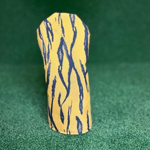 Puede incluir: Una funda para la cabeza del palo de golf con un fondo amarillo y rayas de tigre negras. La funda est&aacute; hecha de un material suave y est&aacute; dise&ntilde;ada para proteger la cabeza del palo de golf. La funda est&aacute; sobre una superficie verde.