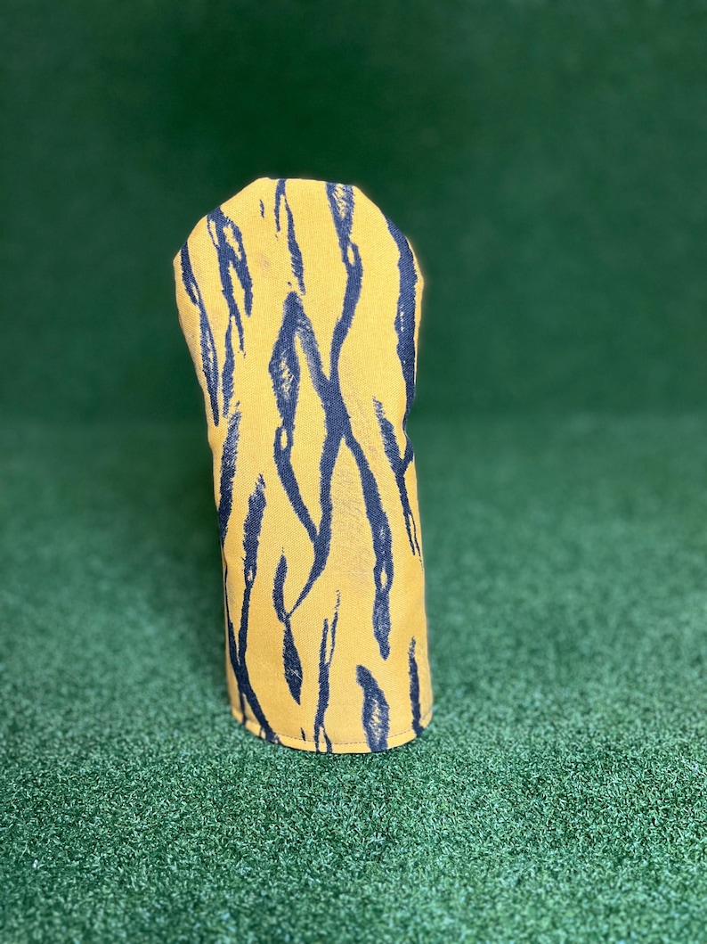 Puede incluir: Una funda para la cabeza del palo de golf con un fondo amarillo y rayas de tigre negras. La funda est&aacute; hecha de un material suave y est&aacute; dise&ntilde;ada para proteger la cabeza del palo de golf. La funda est&aacute; sobre una superficie verde.