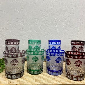 May include: Eight vintage glass tumblers with etched floral designs. The tumblers are in four colors: purple, green, blue, and red. The tumblers are arranged in two rows of four on a woven bamboo surface.