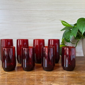 May include: A set of eight stemless drinking glasses in a deep red hue. The glasses are cylindrical with a slightly tapered base, arranged on a wooden surface. A green leafy plant is visible in the background, adding a natural element to the scene.