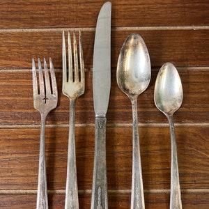 Puede incluir: Un juego de cinco cubiertos de plata plateada, que incluye un cuchillo, un tenedor y tres cucharas, dispuestos sobre una superficie de madera.