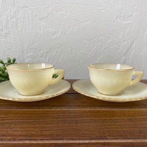 May include: Two white teacups with gold trim and matching saucers on a wooden surface.