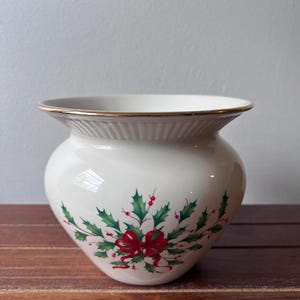 Peut inclure: Un vase en céramique blanc avec un bord doré et un motif festif de houx et de ruban. Le vase a un corps rond et une ouverture légèrement évasée. Le motif de houx comprend des feuilles vertes, des baies rouges et un nœud rouge.