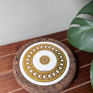 Puede incluir: Posavasos redondo de corcho con una parte superior de cerámica blanca con un diseño circular dorado y marrón. El posavasos tiene un patrón decorativo con un motivo floral central y un borde de formas ovaladas. El posavasos está sobre una superficie de madera.