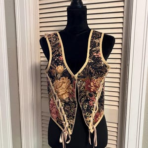 Pode incluir: Um colete sem mangas com base de tecido preto e padrão floral em tons de rosa, amarelo e verde. A peça apresenta um decote em V profundo, acabamento em cor clara e ilhós metálicos frontais com atacadores de fita clara.