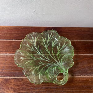 Puede incluir: Un plato de vidrio translúcido de color verde claro con forma de hoja, superficie texturizada y un pequeño asa circular en la base. El plato presenta intrincados patrones de venas en relieve que irradian desde el centro hacia los bordes festoneados.
