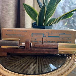 Puede incluir: Un cascanueces Texas Native Inertia vintage, con una base de madera y componentes de latón y metal. La caja original es visible, con el texto "Produces Only Whole Kernels."
