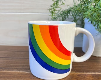 May include: White ceramic mug with a rainbow design. The rainbow is a half circle with red, orange, yellow, green, blue, and indigo stripes.