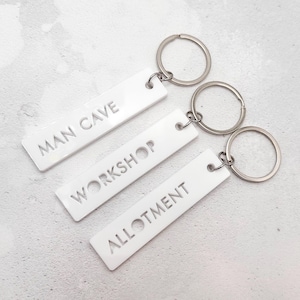 May include: Three white rectangular keyrings with silver rings. Each keyring has a different word engraved: "MAN CAVE", "WORKSHOP", and "ALLOTMENT". The keyrings are made of a smooth, solid material.