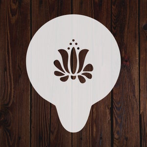May include: White plastic stencil with a floral design. The design features a stylized flower with three petals and a central stem.