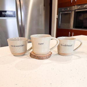 May include: Three speckled ceramic mugs with a light beige finish and a natural clay base. Each mug features a single word printed in black lowercase letters: cockwomble, cunt, and knobhead. The centre mug rests on a small, circular wooden slice.