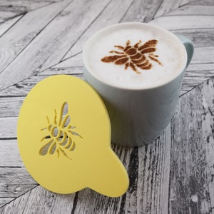 Könnte beinhalten: Eine gelbe Kunststoffschablone in Form einer Biene, mit einer weißen Keramiktasse mit einem Bienendesign aus Zimt auf dem Schaum.