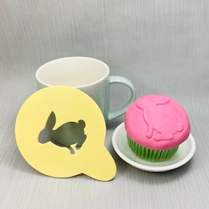 May include: A light green mug, a yellow bunny stencil, and a cupcake with pink icing and a bunny design are arranged on a light grey surface. The cupcake sits on a small white plate with green icing.