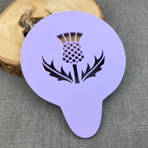 May include: Purple plastic stencil of a thistle flower with a detailed design. The stencil is shaped like a circle with a pointed bottom.