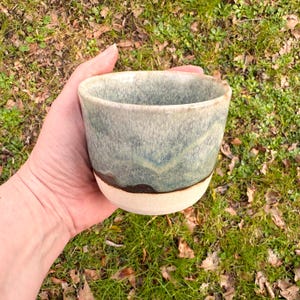 May include: A ceramic cup with a wide, open top and a rounded base. The upper portion features a mottled, blended glaze in shades of sage green, blue, and grey, while the lower section remains an unglazed, matte off-white or cream-coloured clay.