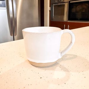 May include: A white ceramic mug with a rounded body and a large handle. The base of the mug is a natural, unglazed tan colour. The mug is on a speckled countertop.