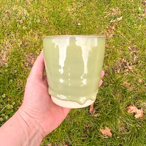 May include: A handmade ceramic cup with a pale green glaze. The cup has a rounded shape with a white base and a glossy finish. The glaze drips down the sides, creating a unique texture. The cup is held against a background of green grass.