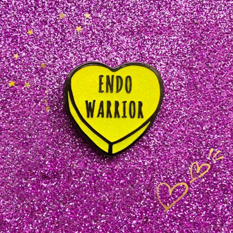 May include: A yellow heart-shaped enamel pin with the words "ENDO WARRIOR" in black lettering. The pin has a black border and is set against a sparkly purple background. Gold heart illustrations are in the lower right corner.