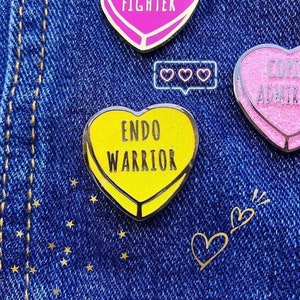 May include: A close-up of several heart-shaped enamel pins on a blue denim background. The pins have text such as "ENDO WARRIOR" and "COPE ADMIRABLY". The pin in the center is yellow with black text. Other pins are pink and white.