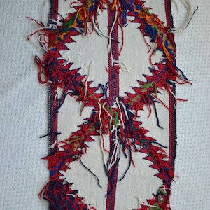 May include: A white woven fabric with a red, blue, green, and yellow geometric pattern. The fabric has fringe along the edges.