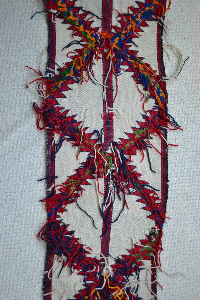 May include: A white woven fabric with a red, blue, green, and yellow geometric pattern. The fabric has fringe along the edges.