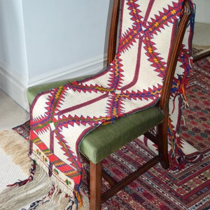 May include: A white chair with a green cushion is draped with a colorful woven throw with fringe. The throw has a geometric pattern in red, blue, yellow, and white. The chair is sitting on a patterned rug.
