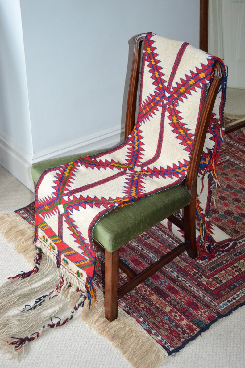May include: A white chair with a green cushion is draped with a colorful woven throw with fringe. The throw has a geometric pattern in red, blue, yellow, and white. The chair is sitting on a patterned rug.