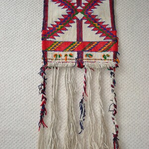 May include: A white woven tapestry with red, blue, green, and yellow geometric patterns. The tapestry has long white fringe with red, blue, and white accents.