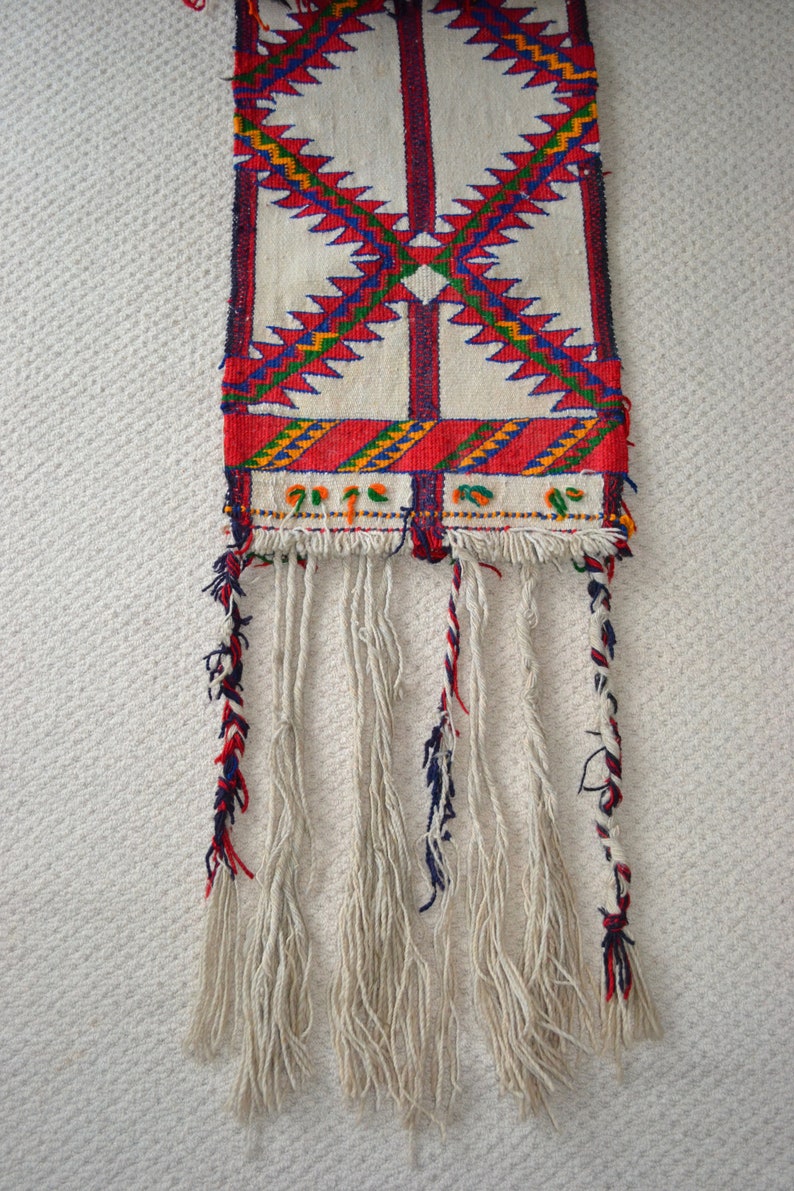 May include: A white woven tapestry with red, blue, green, and yellow geometric patterns. The tapestry has long white fringe with red, blue, and white accents.