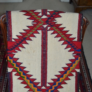 May include: A white woven rug with a geometric pattern in red, blue, green, and orange. The pattern features a series of triangles and zigzags.