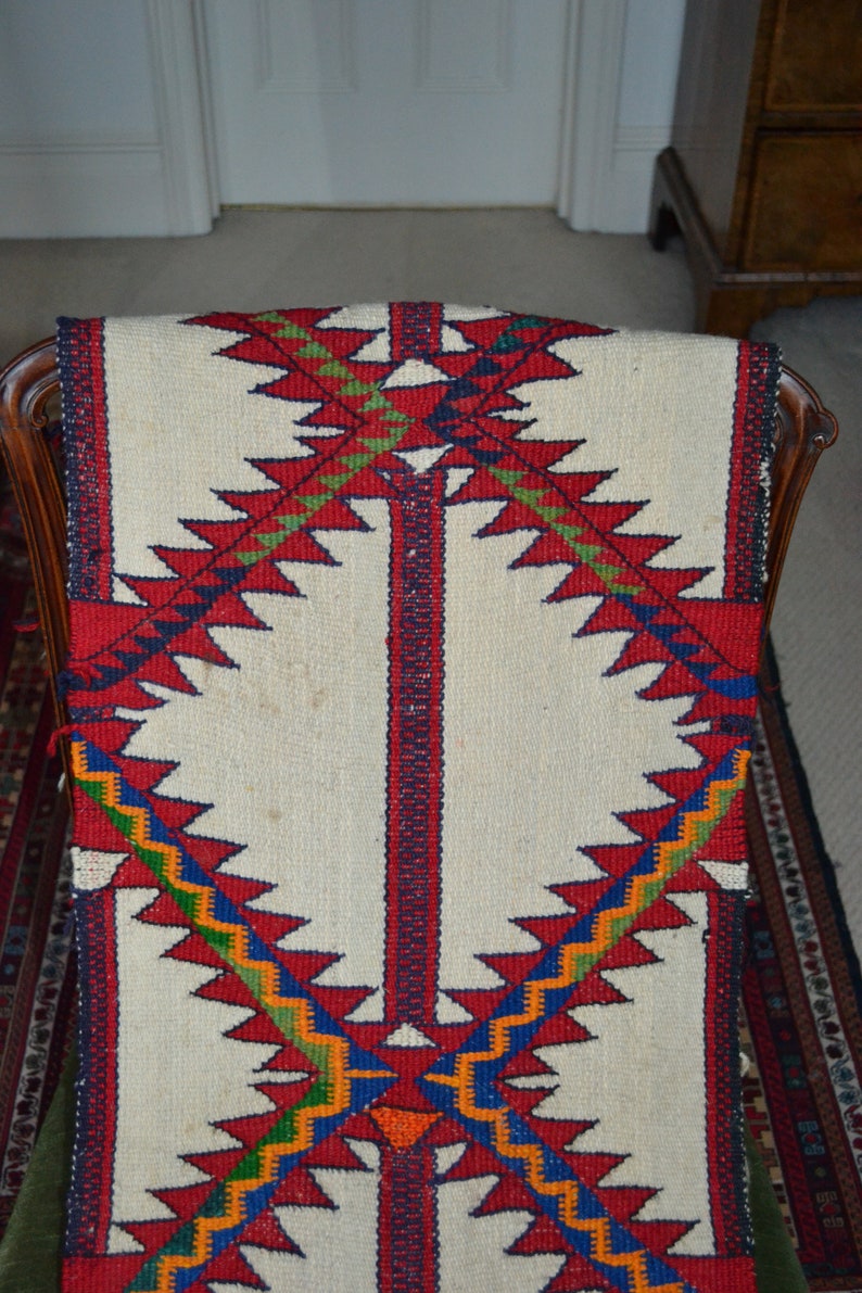 May include: A white woven rug with a geometric pattern in red, blue, green, and orange. The pattern features a series of triangles and zigzags.