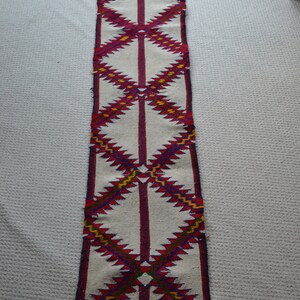 May include: A long, white runner rug with a geometric pattern in red, blue, yellow, and green. The rug has a fringe on the bottom.
