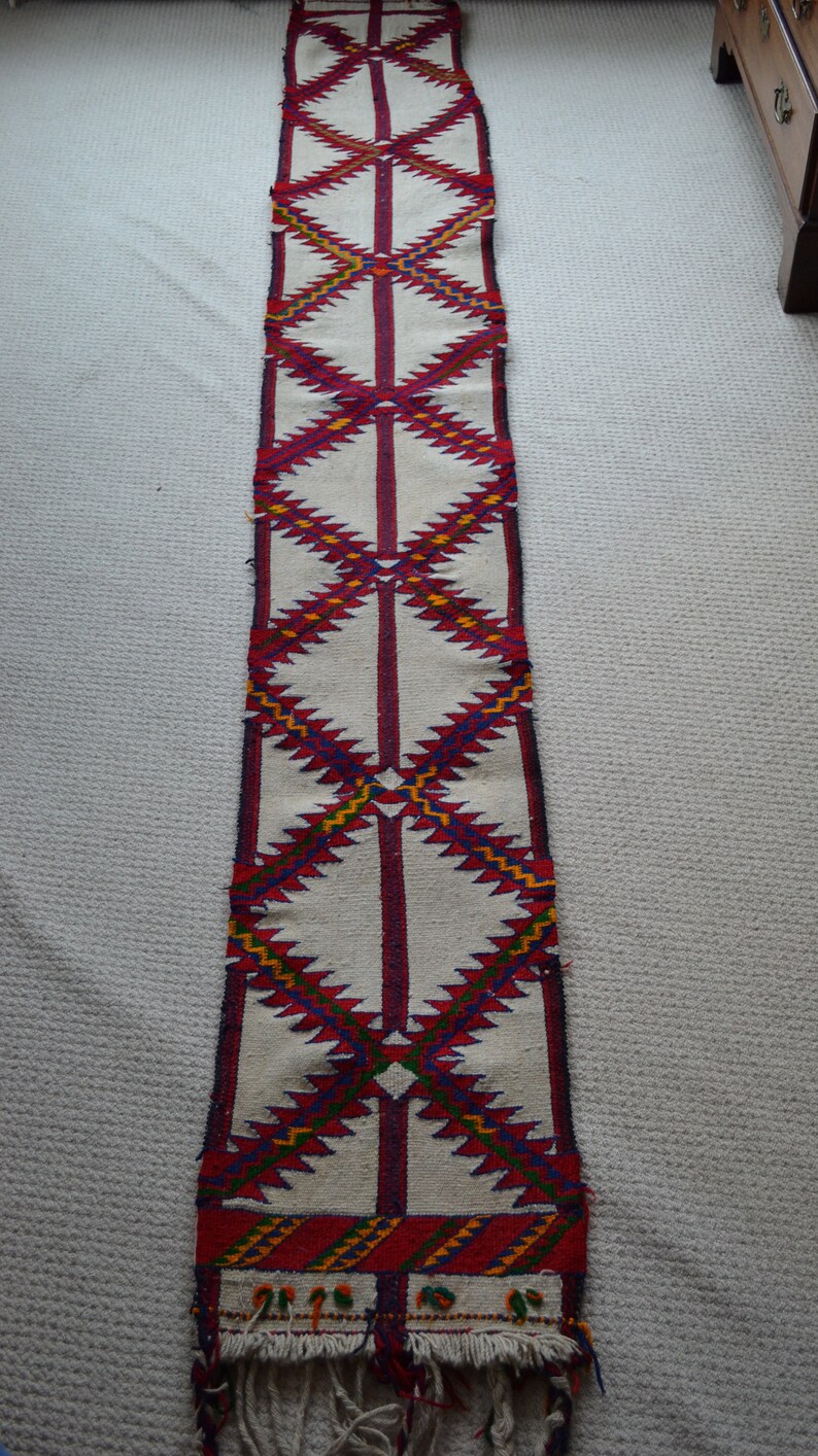 May include: A long, white runner rug with a geometric pattern in red, blue, yellow, and green. The rug has a fringe on the bottom.