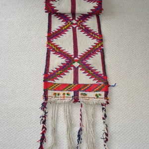 May include: A white woven rug with a geometric pattern in red, blue, and yellow. The rug has a fringe on the bottom.