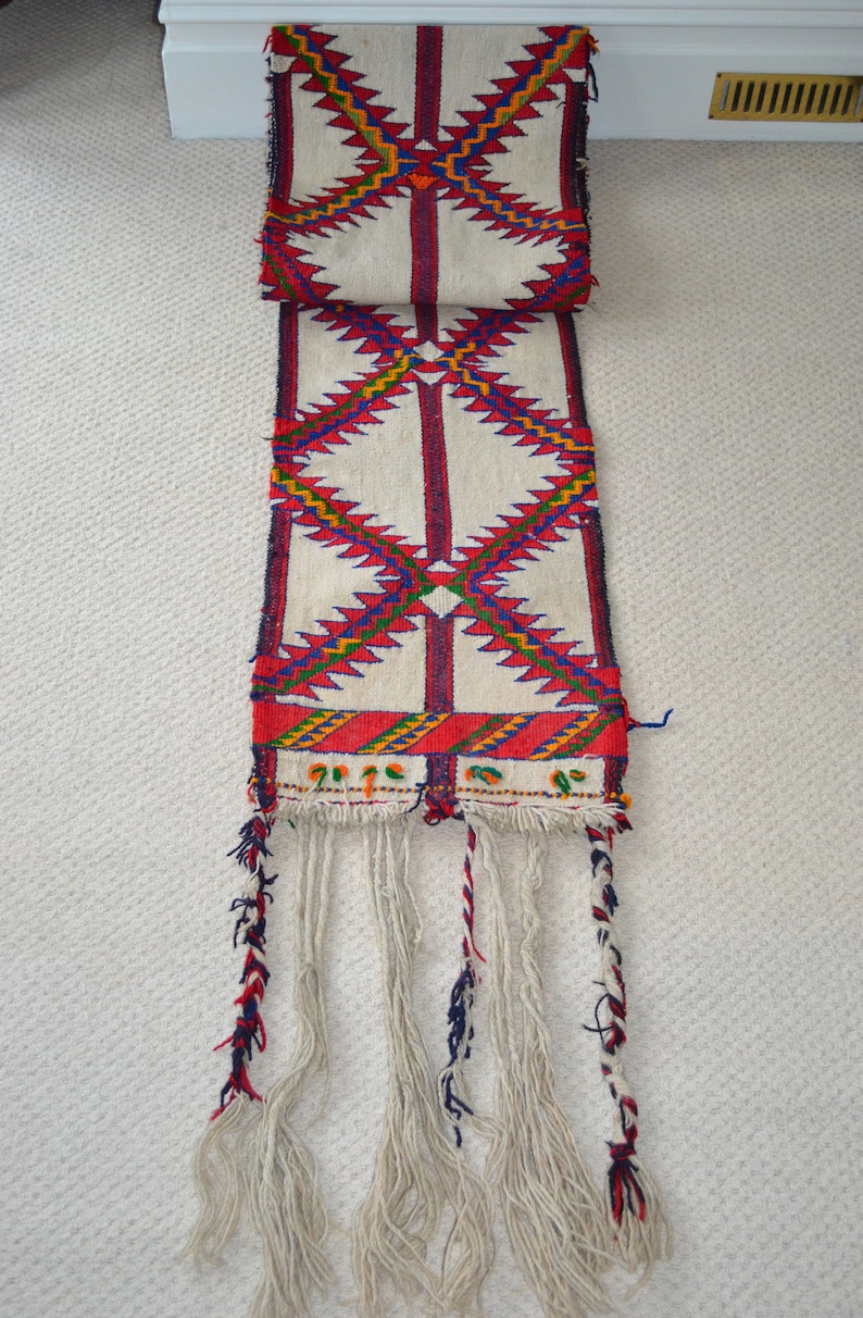 May include: A white woven rug with a geometric pattern in red, blue, and yellow. The rug has a fringe on the bottom.
