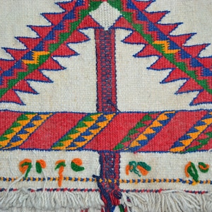 May include: A white woven rug with a geometric pattern in red, blue, green, and yellow. The rug has a fringe border.