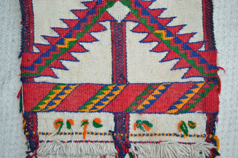 May include: A white woven rug with a geometric pattern in red, blue, green, and yellow. The rug has a fringe border.