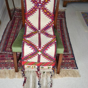 May include: A white woven rug with red, blue, and yellow geometric patterns. The rug has long fringe tassels and is draped over a wooden chair.