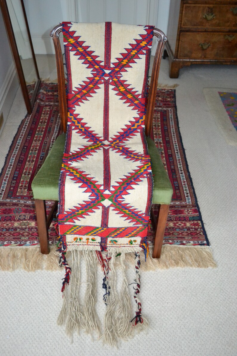 May include: A white woven rug with red, blue, and yellow geometric patterns. The rug has long fringe tassels and is draped over a wooden chair.