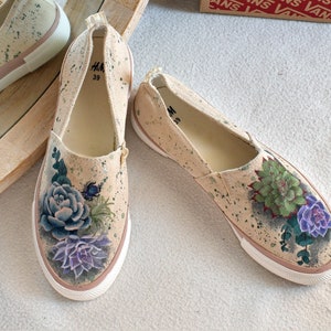 May include: A pair of beige canvas slip-on shoes with a floral design. The shoes have a white sole and are decorated with green and purple succulents.