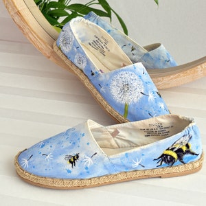 May include: A pair of blue canvas espadrilles with a dandelion and bee design. The shoes have a light blue background with white dandelion seeds and a yellow and black bee.