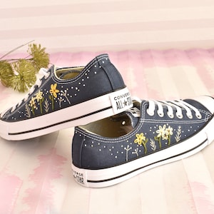 Peut inclure: Une paire de baskets en toile bleu marine avec des lacets blancs et une semelle en caoutchouc blanche. Les baskets sont décorées de fleurs jaunes brodées et de tiges vertes. Le texte "Converse All Star" est visible sur le côté d'une basket.