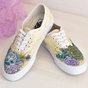 Könnte beinhalten: Ein Paar weiße Canvas-Sneaker mit einem gelben Hintergrund aus Farbspritzern. Die Sneaker sind handbemalt mit einem floralen Design, das Sukkulenten in Grün-, Lila- und Blautönen zeigt.